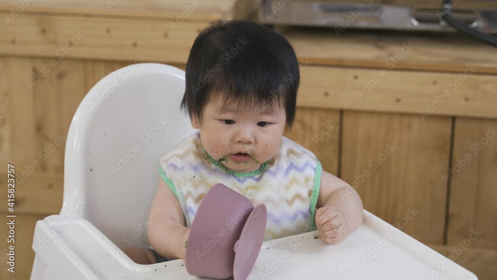 portrait adorable young child is looking at the overturned container with curiosity on a baby booster seat with food spills at home. portrait adorable young child is looking at the overturned container with curiosity on a baby booster seat with food spills at home.