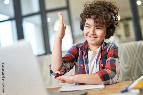 Learning is fun. Curious latin american little boy wearing headphones, using laptop while studying at home. School kid raised his hand during online lesson