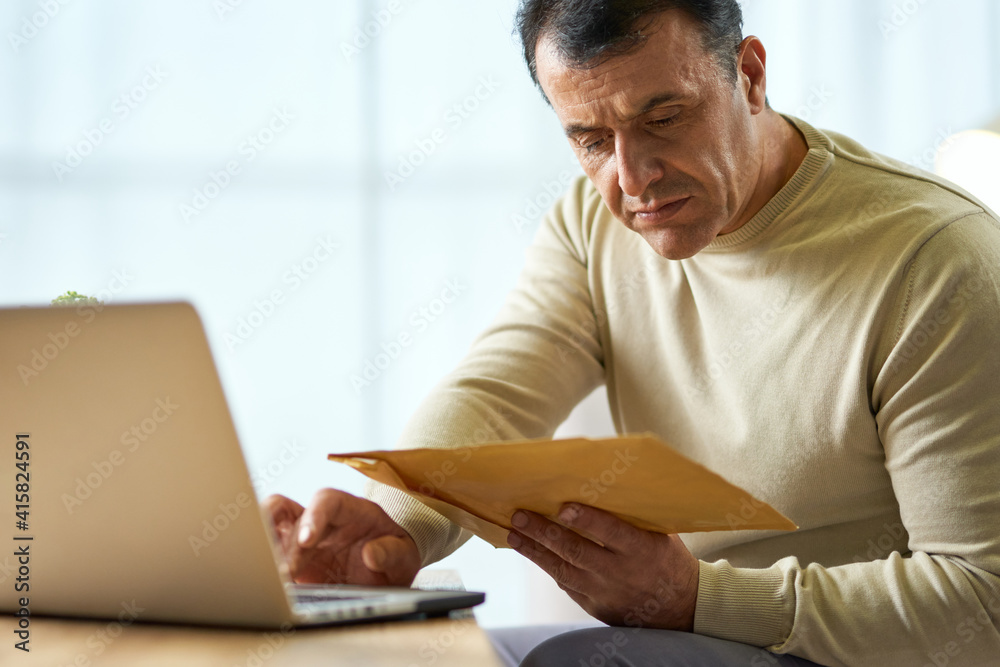 Portrait of busy latin middle aged businessman looking at the envelope, using laptop while working from home Portrait of busy latin middle aged businessman looking at the envelope, using laptop while working from home