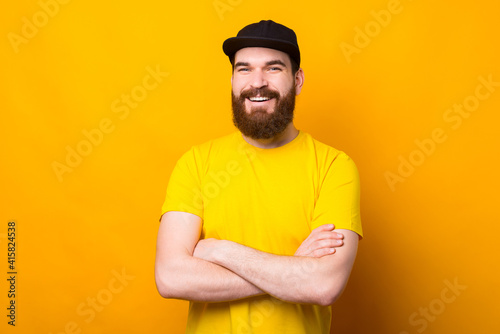 Happy smiling bearded hipster man in yellow shirt standing with crossed arms.