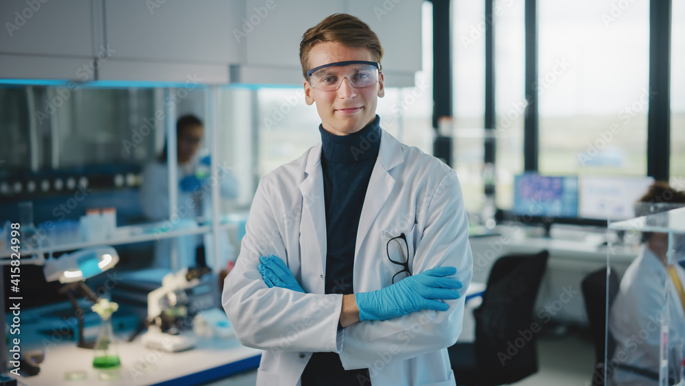 Medical Science Laboratory: Handsome Young Scientist Wearing White Coat and Safety Glasses, Posing and Smiles Looking at Camera with Crosses Arms. Diverse Team of Specialists. Medium Portrait Shot.