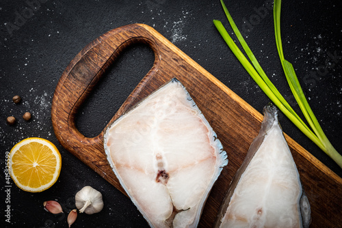Frozen raw fish steak on wooden cutting board with spices on black background.