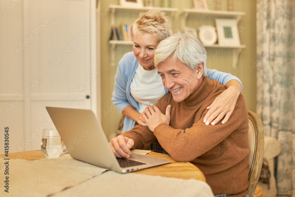 Indoor shot of handsome senior man and his attractive wife using generic laptop together at home enjoying online communication, talking to family via video conference call. smiling happily