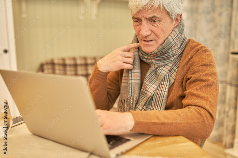 Unhealthy frustrated mature man with gray hair sitting at desk wrapped in scarf having sore throat, feeling sick, trying to consecrate on working task, using wireless internet connection on laptop Unhealthy frustrated mature man with gray hair sitting at desk wrapped in scarf having sore throat, feeling sick, trying to consecrate on working task, using wireless internet connection on laptop
