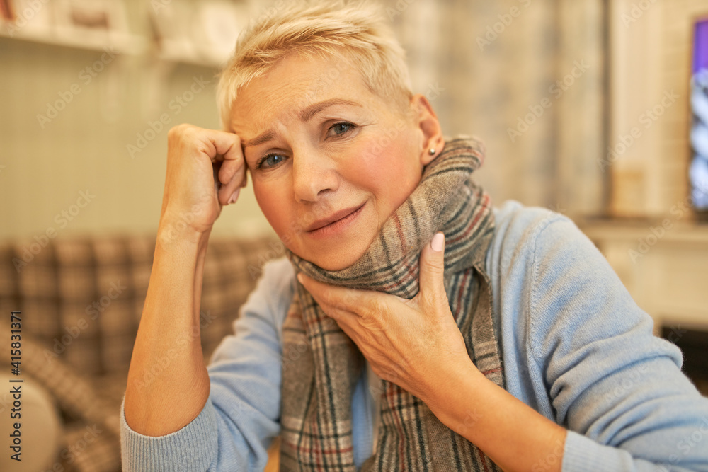 Infection, virus, disease and sickness concept. Indoor image of desperate sad mature woman wearing warm scarf suffering from sore throat, migraine and fever, frowning, having mournful look Infection, virus, disease and sickness concept. Indoor image of desperate sad mature woman wearing warm scarf suffering from sore throat, migraine and fever, frowning, having mournful look