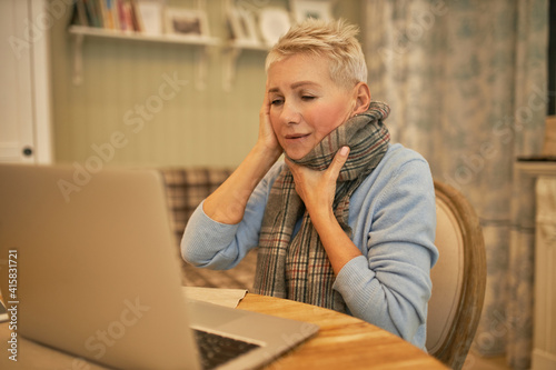 Frustrated mournful retired woman with terrible headache and sore throat sitting in front of open laptop at home complaining of contagious disease symptoms to doctor during online distant consultation