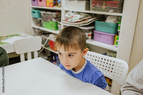 Young cute boy with down syndrome in blue shirt sitting at white desk and studying. Copy space.