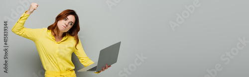 irritated woman holding laptop while standing with clenched fist on grey, banner