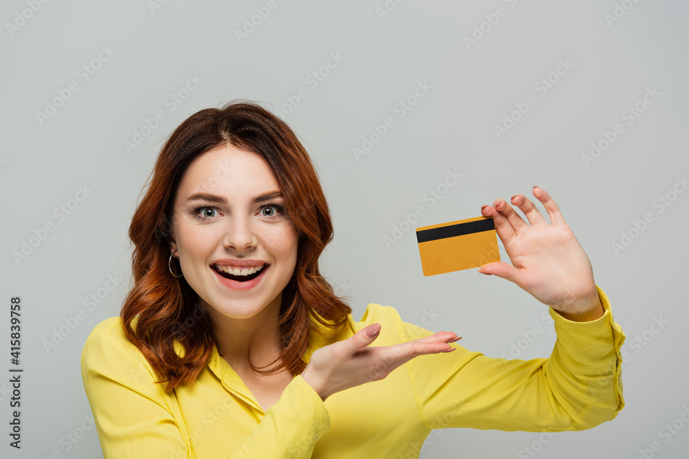 cheerful woman looking at camera while pointing at credit card isolated on grey cheerful woman looking at camera while pointing at credit card isolated on grey
