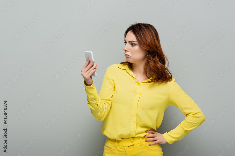 shocked woman looking at mobile phone while standing with hand on hip on grey shocked woman looking at mobile phone while standing with hand on hip on grey