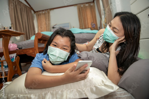 Asian mother and Kid wear mask and using mobile phone together. Mother and Daughter lie on the bed and learn online study at home. Happy family in New normal Lifestyle concept.