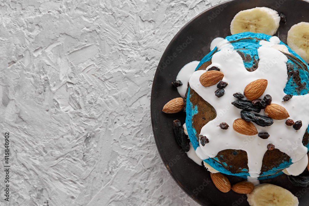 Vegan blue pancakes with sour cream and banana slices on a plate sprinkled with almonds, raisins on a gray textured background. Top view with space to copy text