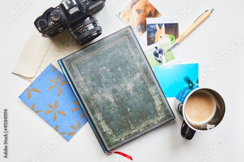 A beautiful notebook surrounded by a classical camera, some photos, a postcard, a pen and a coffee.