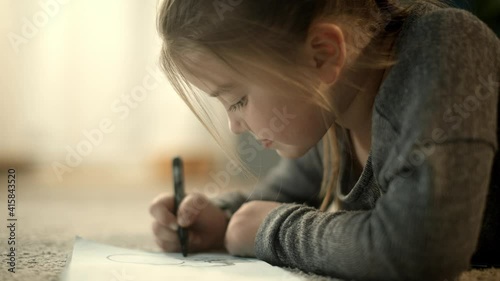 Little, happy preschool girl drawing with a marker in her notebook, smiling and laughing laying on the carpet. Artistic children and apartment concept.