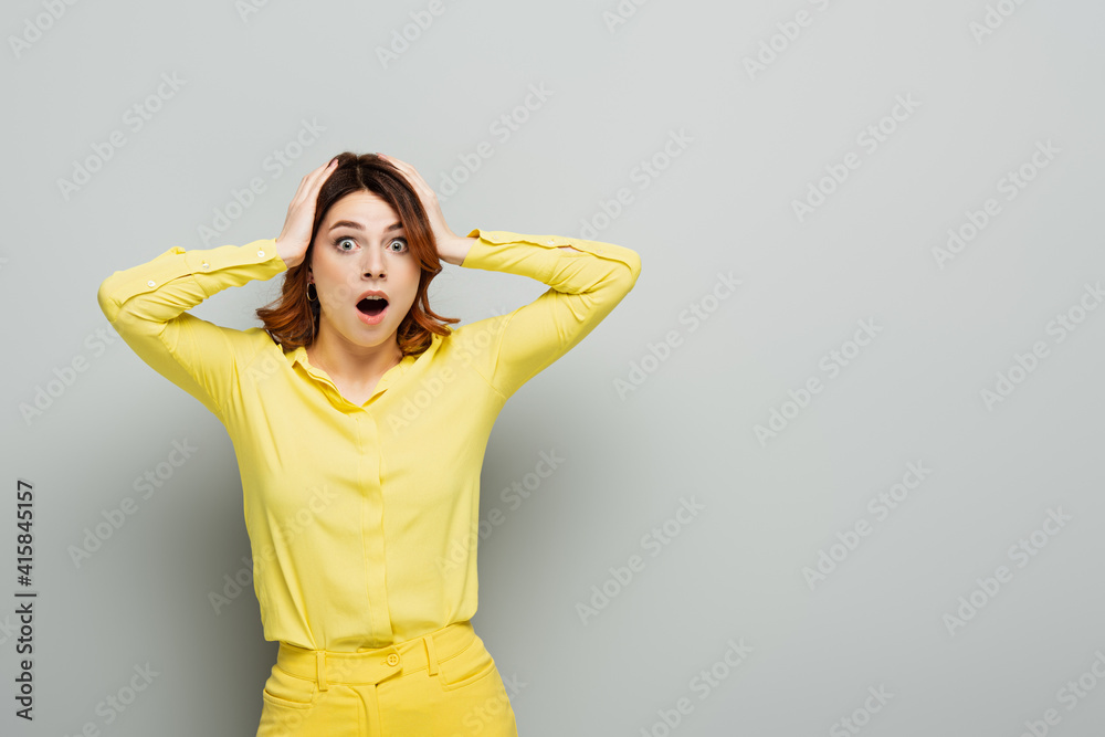 shocked woman touching head while standing with open mouth on grey shocked woman touching head while standing with open mouth on grey