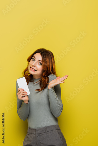 фотографія clueless young woman with curly hair holding smartphone and gesturing on yellow