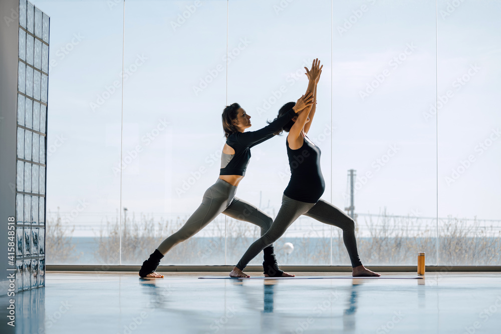 Young female yoga instructor teaching yoga in studio to a pregnant woman. Prenatal practice, working out indoor in a big gym, fitness center. Teacher helping to master, full length image.