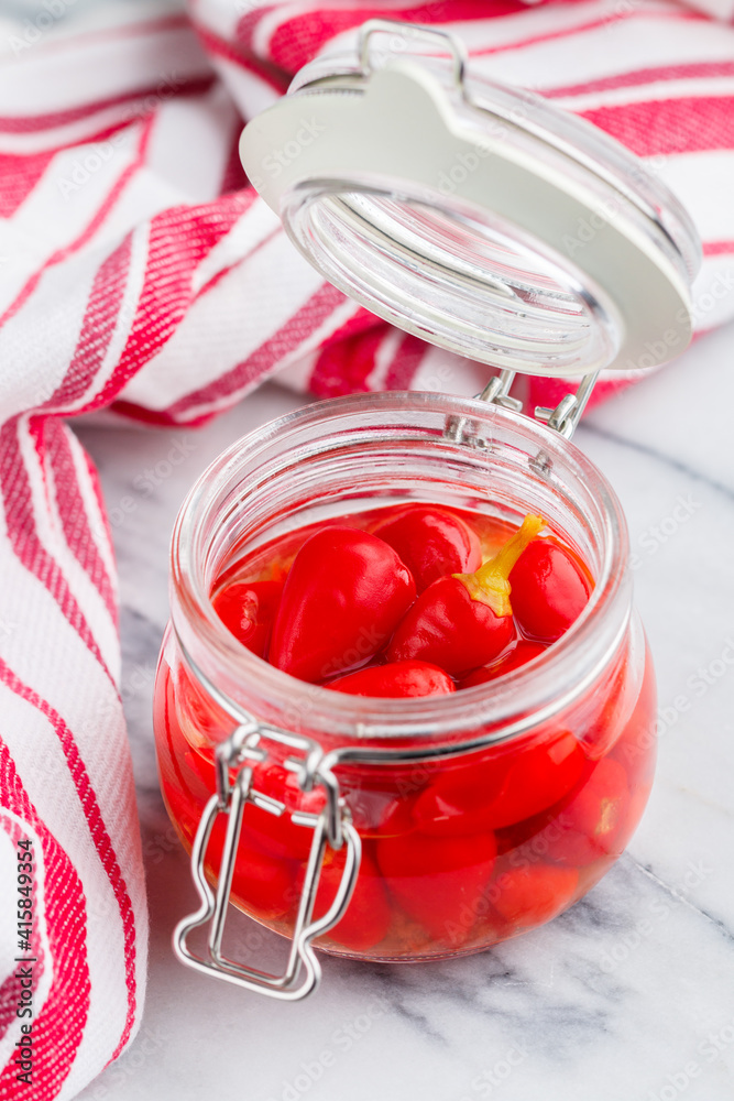 Red hot chili pepper. Piri piri. Pickled peppers in a glass jar on a marble table. A gourmet snack for meat and poultry. (Capsicum chinense). Selective Focus Red hot chili pepper. Piri piri. Pickled peppers in a glass jar on a marble table. A gourmet snack for meat and poultry. (Capsicum chinense). Selective Focus