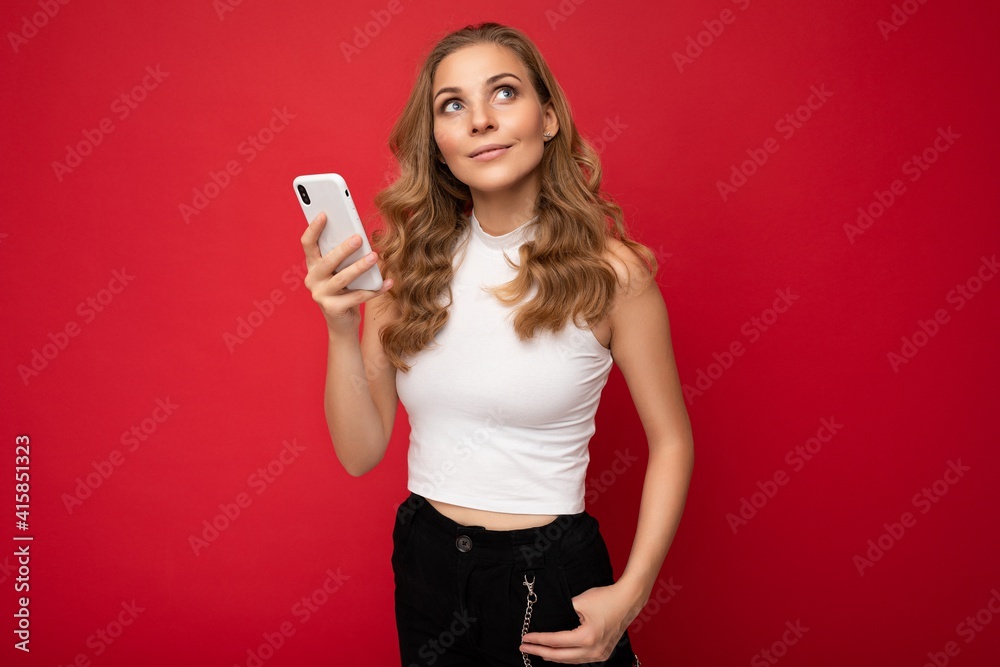 happy pretty young blonde woman wearing white t-shirt isolated over red background using smartphone and chatting via mobile phone looking to the side and thinking happy pretty young blonde woman wearing white t-shirt isolated over red background using smartphone and chatting via mobile phone looking to the side and thinking