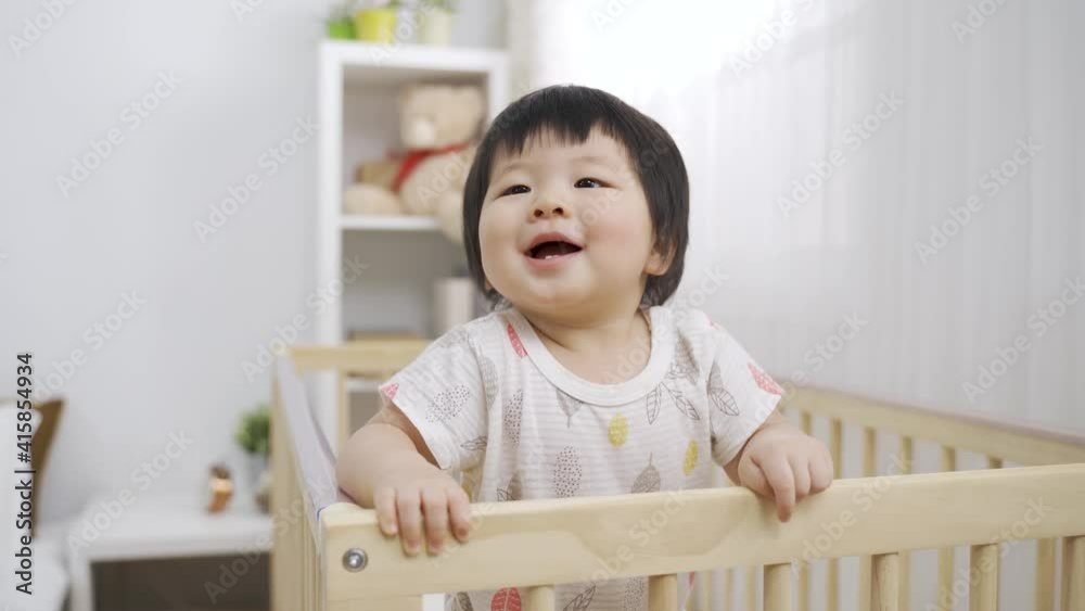 tilt up adorable asian baby is smiling at the distance and babbling while leaning on the wooden crib playpen in the bedroom at home. tilt up adorable asian baby is smiling at the distance and babbling while leaning on the wooden crib playpen in the bedroom at home.