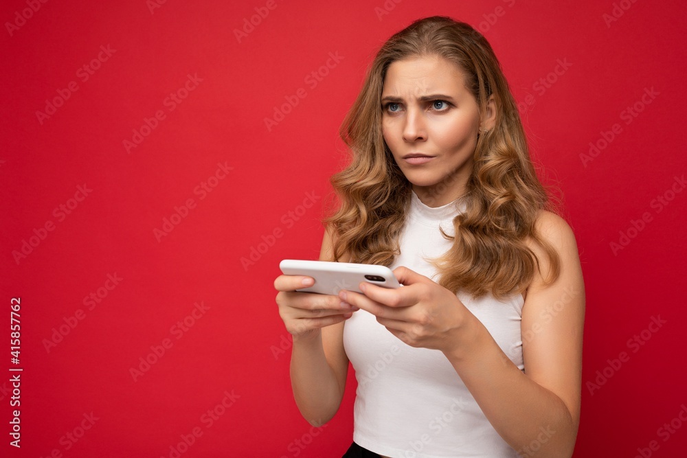 Serious beautiful young blonde woman wearing white t-shirt isolated over red background using smartphone and texting message via mobile phone looking forward Serious beautiful young blonde woman wearing white t-shirt isolated over red background using smartphone and texting message via mobile phone looking forward