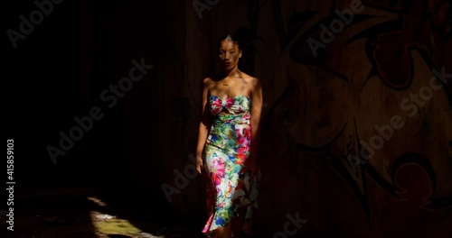 A sexy girl in a long dress walks out from a dark spot with grafitti on the wall into the sun rays photo
