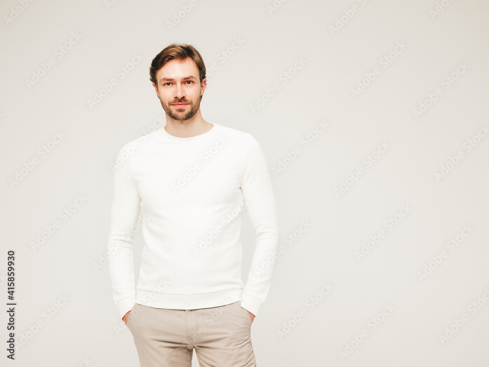 Portrait of handsome smiling hipster lumbersexual businessman model wearing casual white sweater and trousers. Fashion stylish man posing against gray background in studio
