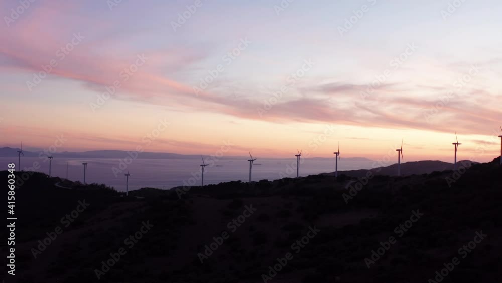Aerial wide shot showing silhouette of wind farm turbines during sunset on top of hill over the cloudscape. Aerial wide shot showing silhouette of wind farm turbines during sunset on top of hill over the cloudscape.