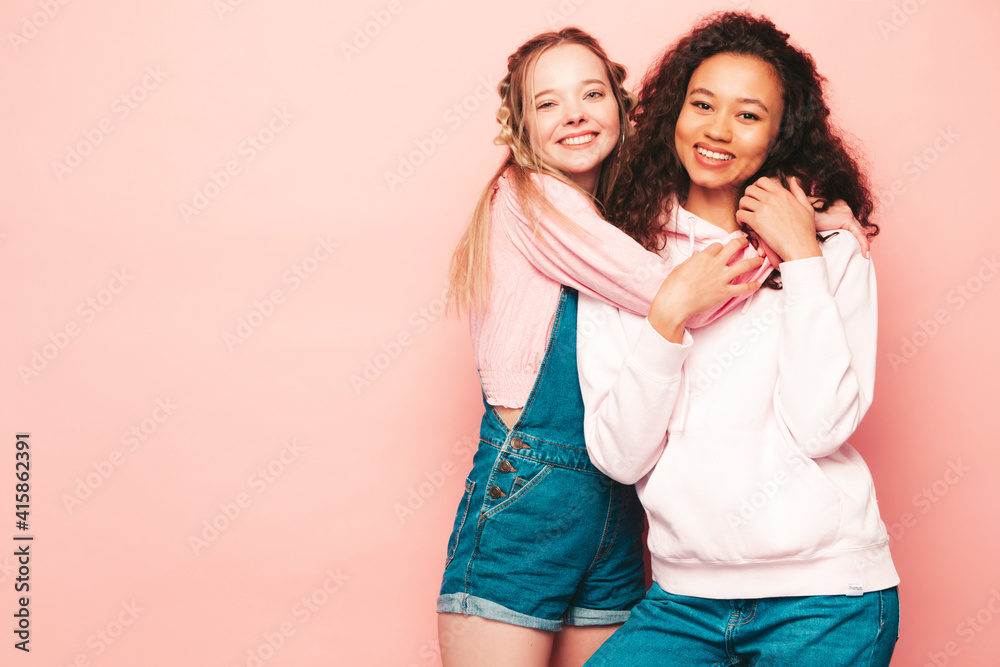 Two young beautiful smiling international hipster female in trendy summer clothes. Sexy carefree women posing near pink wall in studio. Positive models having fun. Concept of friendship Two young beautiful smiling international hipster female in trendy summer clothes. Sexy carefree women posing near pink wall in studio. Positive models having fun. Concept of friendship