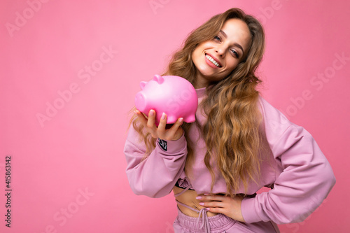 Portrait photo of happy positive smiling young beautiful attractive woman with wavy long blonde hair with sincere emotions wearing trendy pink hoodie isolated over pink background with copy space and