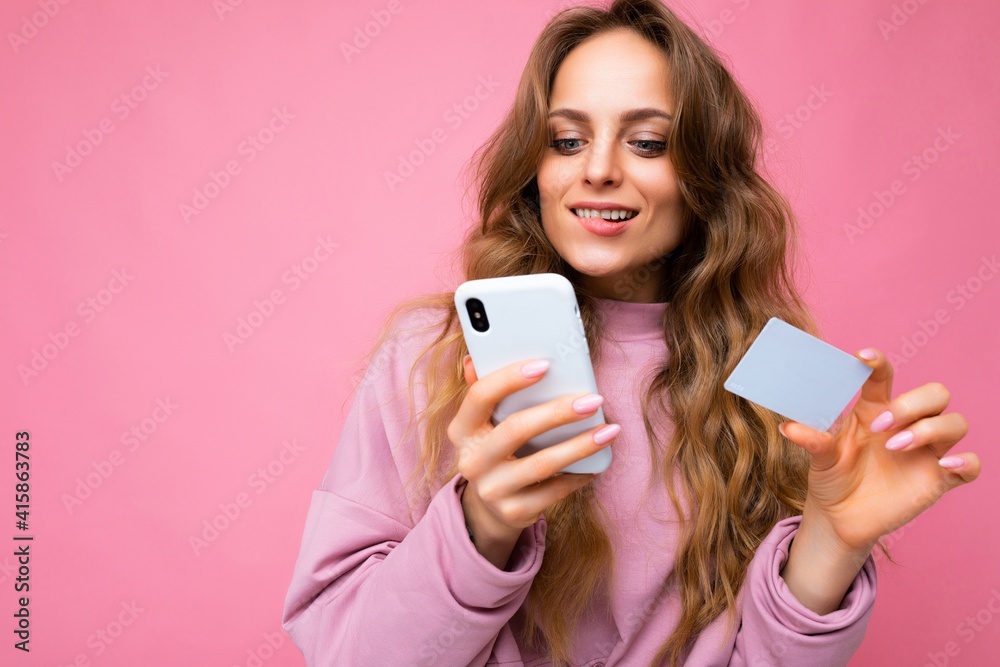 Beautiful sexy positive young blonde curly woman wearing pink clothes isolated over pink background using mobile phone paying online shopping through credit card looking at smartphone display Beautiful sexy positive young blonde curly woman wearing pink clothes isolated over pink background using mobile phone paying online shopping through credit card looking at smartphone display