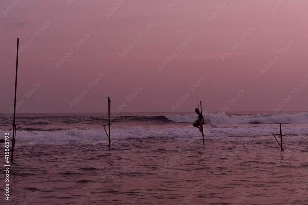 Tradycyjni rybacy wędkarze podczas połowy na palach w oceanie, na tle zachodzącego słońca.