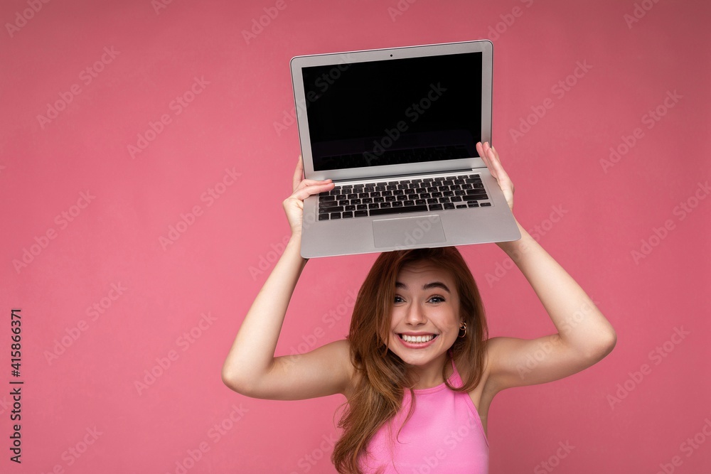 Naklejka premium Photo of happy satisfied Beautiful young blond woman holding computer laptop with empty monitor display wearing pink crop top looking at camera isolated on pink background