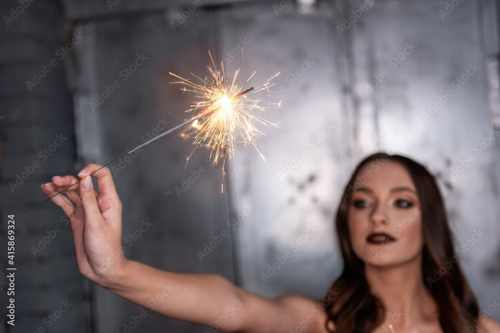 Obraz premium Blurred Beautiful girl, holds bright sparklers in focus in hands. With professional evening make-up, in a gold top, against a gray metal wall with green monsteras background. Holiday party. Copy paste