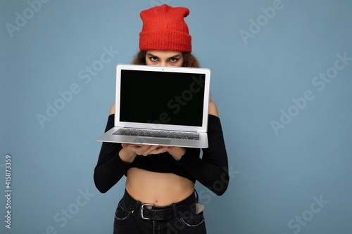 Close-up portrait of funny beautiful dark blond woman holding laptop computer looking at camera wearing black crop top and red and orange do-rag isolated over light blue wall background photo