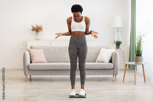 Displeased Black Woman Standing On Scales Weighing Herself At Home