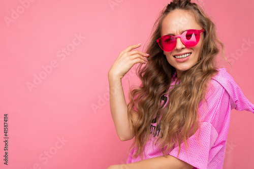 Closeup of attractive smiling happy young blonde woman wearing everyday stylish clothes and modern sunglasses isolated on colorful background wall looking at camera