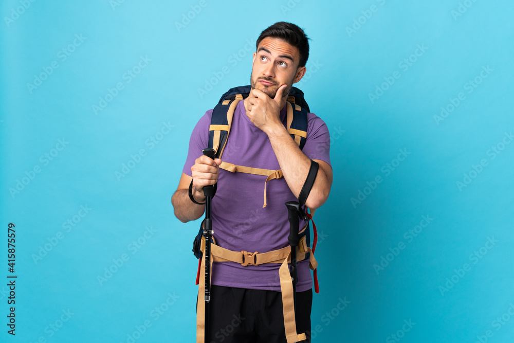 Young caucasian man with backpack and trekking poles isolated on blue background having doubts