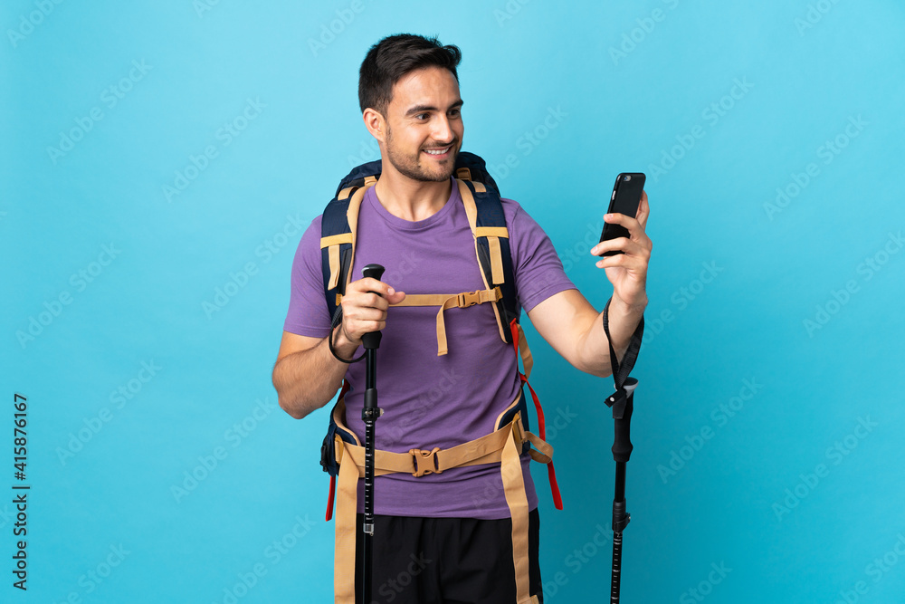 Young caucasian man with backpack and trekking poles isolated on blue background making a selfie