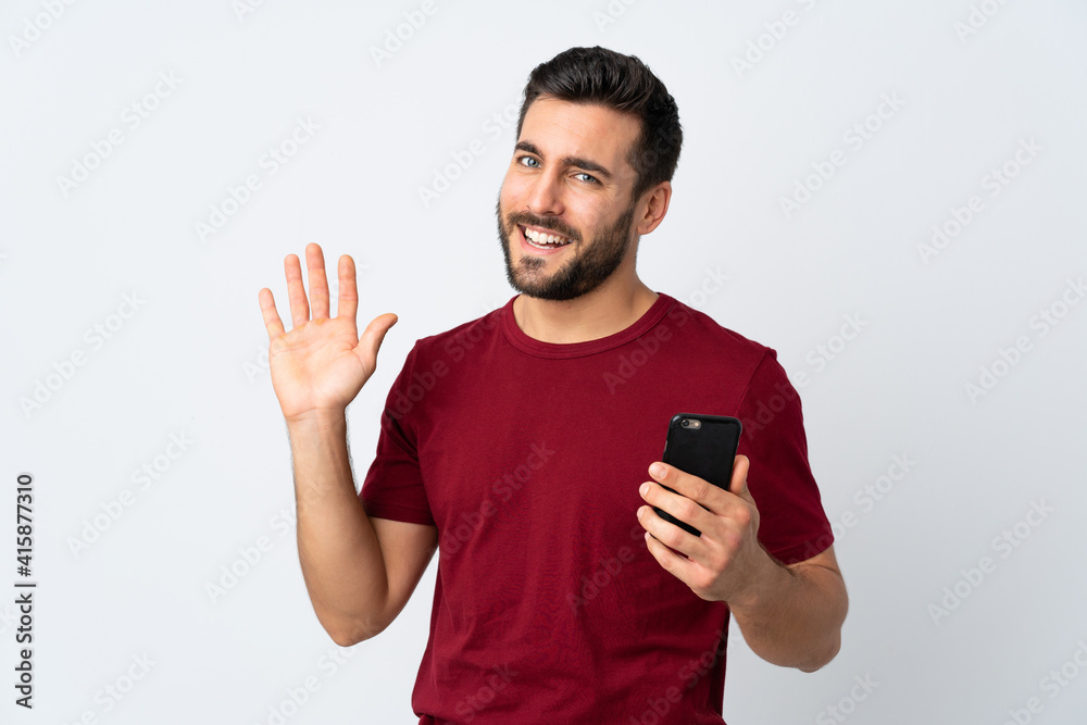 Young handsome man with beard using mobile phone isolated on white background saluting with hand with happy expression Young handsome man with beard using mobile phone isolated on white background saluting with hand with happy expression