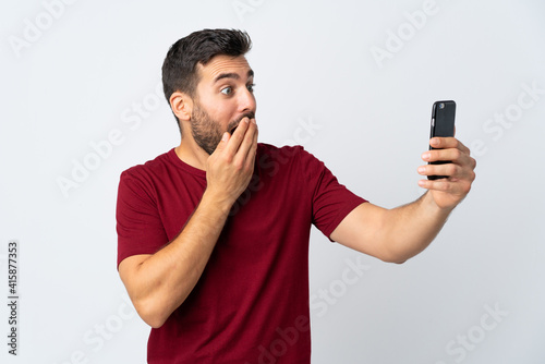 Young handsome man with beard using mobile phone isolated on white background with surprise and shocked facial expression © luismolinero