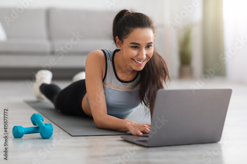 Sporty woman exercising with dumbbells at home, using laptop