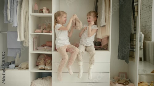 Family wardrobe. Happy twin sisters on shelve in closet with natural organic clothes in cozy dressing room play joyfully