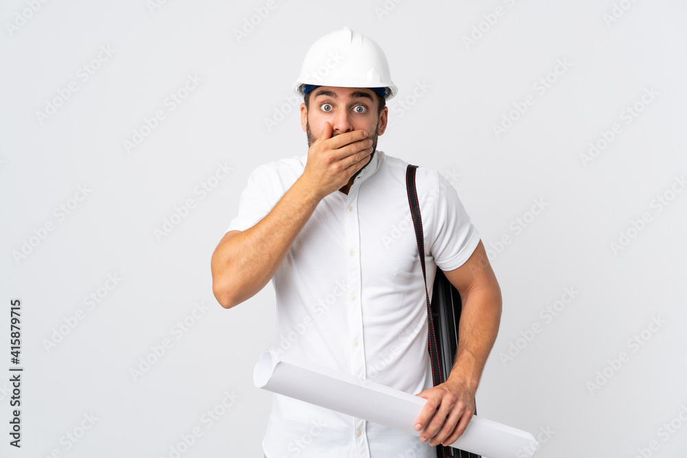 Young architect man with helmet and holding blueprints isolated on white background covering mouth with hands Young architect man with helmet and holding blueprints isolated on white background covering mouth with hands