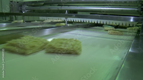 Instant noodle briquettes move along a conveyor belt on an automated line in a pasta factory. The briquettes are moving towards the camera.