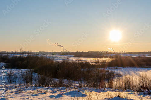 Winter evening landscape with sunset and multicolor sky. Colorful snow with animal footprints illuminated by sunset. On the horizon is an urban industrial zone with smoking chimneys