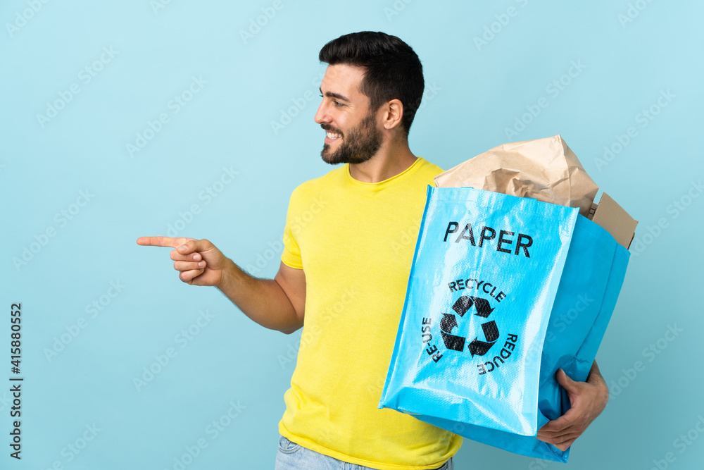 Young caucasian man holding a recycle bag isolated on blue background pointing to the side to present a product Young caucasian man holding a recycle bag isolated on blue background pointing to the side to present a product