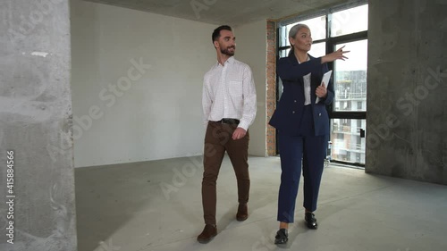 Delighted client listening to female real estate agent while walking around large empty space of apartment for sale. Professional realtor telling about future interior of property to buyer