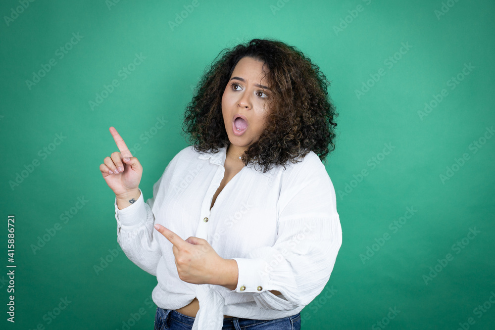 Young african american girl wearing white shirt over green background surprised and pointing her fingers side