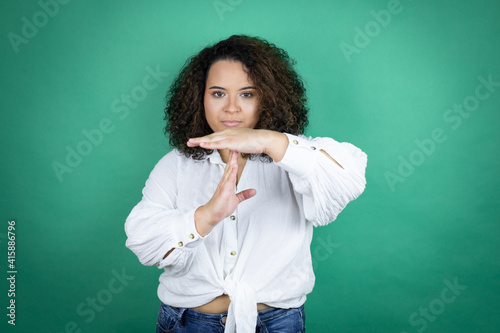 Young african american girl wearing white shirt over green background Doing time out gesture with hands, frustrated and serious face